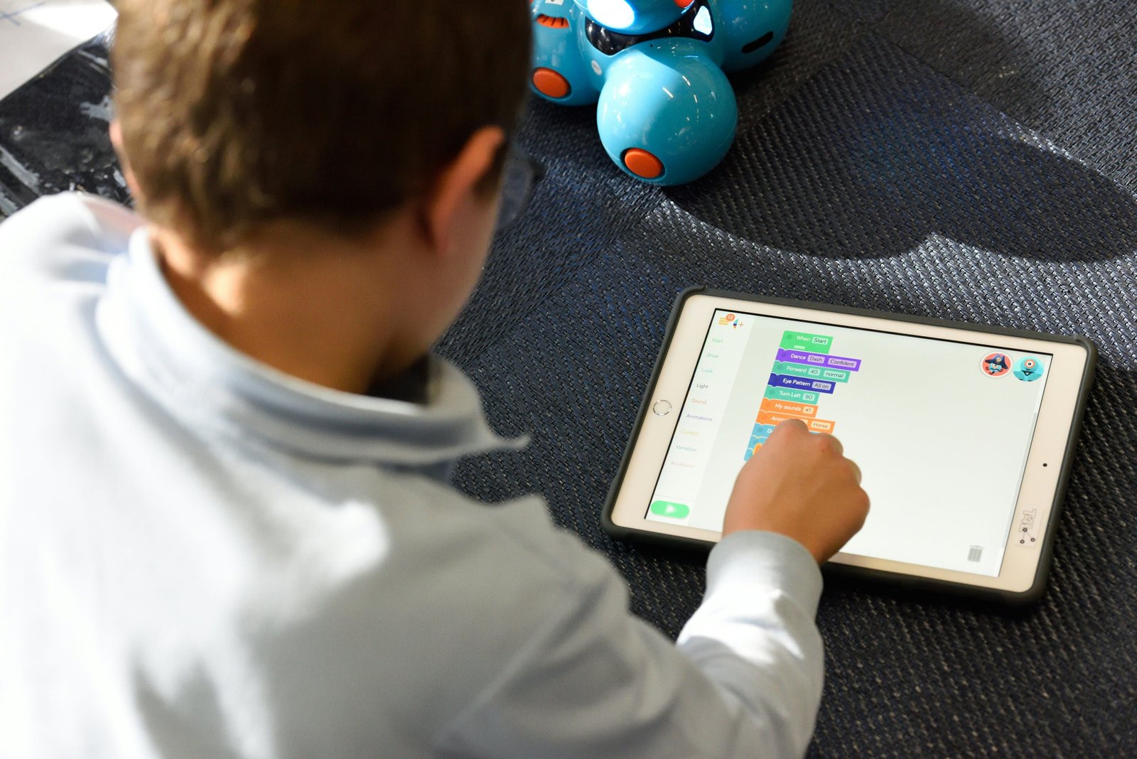 An overhead view of a child in a white long-sleeved shirt using a tablet device displaying what appears to be an educational application, with blue toy robots visible on a dark textured surface beside the tablet.